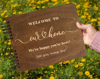 May include: A brown wooden guest book with a spiral binding. The cover has the text "Welcome to our home" with a heart symbol and the text "We're happy you're here! Add your message here!"