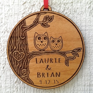 May include: A round wooden ornament with a tree and two owls perched on a branch. The ornament is engraved with the names "Laurie & Brian" and the date "9-12-15". A red ribbon is attached for hanging. The ornament is made of light-colored wood.