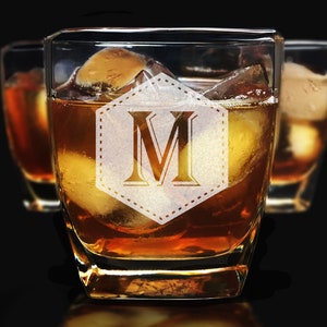 Puede incluir: Vasos de whisky cuadrados llenos de l&iacute;quido &aacute;mbar y cubitos de hielo. El vaso delantero presenta un monograma "M" dentro de una forma decorativa. El fondo muestra dos vasos adicionales, todo sobre un fondo oscuro.