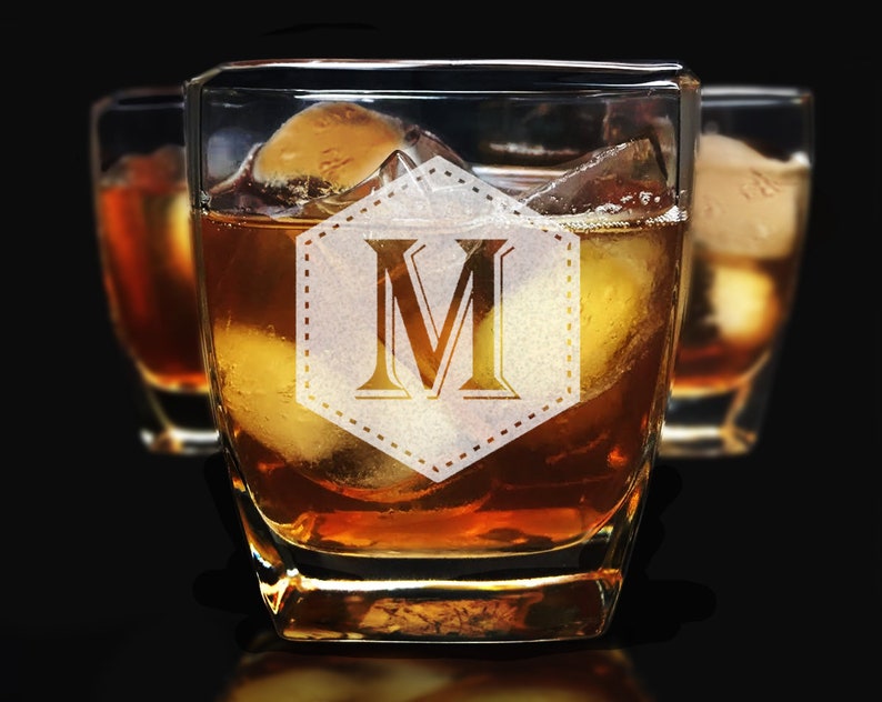 Puede incluir: Vasos de whisky cuadrados llenos de l&iacute;quido &aacute;mbar y cubitos de hielo. El vaso delantero presenta un monograma "M" dentro de una forma decorativa. El fondo muestra dos vasos adicionales, todo sobre un fondo oscuro.