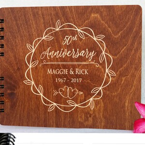 Puede incluir: Un &aacute;lbum de aniversario de madera con encuadernaci&oacute;n en espiral. La portada est&aacute; grabada con "50th Anniversary", nombres y fechas dentro de una corona decorativa. La madera tiene un tono marr&oacute;n c&aacute;lido.