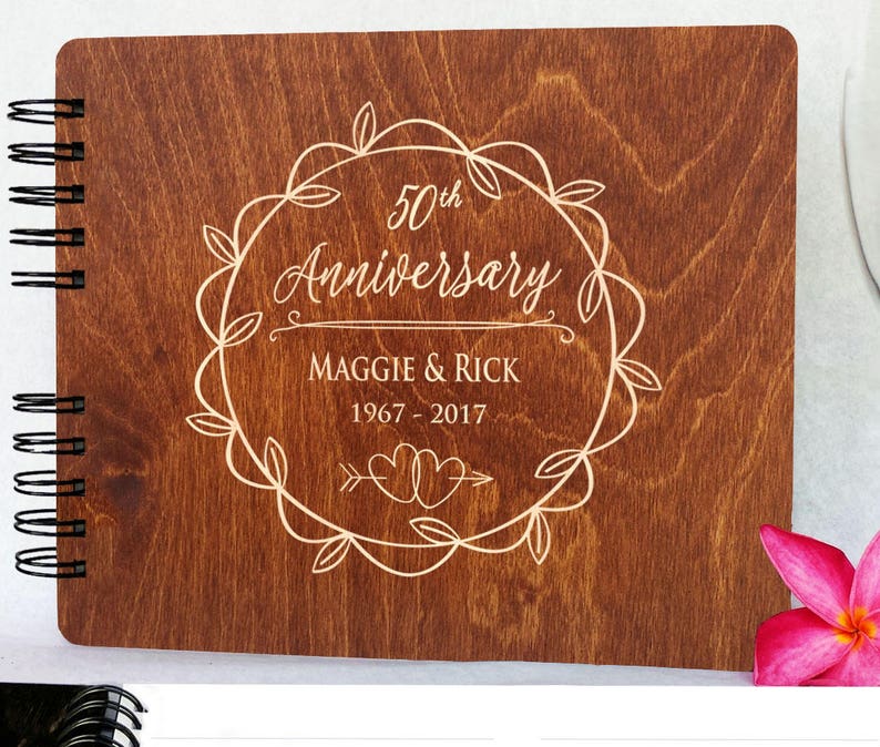 Puede incluir: Un &aacute;lbum de aniversario de madera con encuadernaci&oacute;n en espiral. La portada est&aacute; grabada con "50th Anniversary", nombres y fechas dentro de una corona decorativa. La madera tiene un tono marr&oacute;n c&aacute;lido.