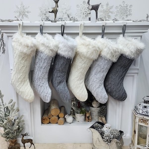 May include: Six gray and white knitted Christmas stockings with faux fur trim hanging on a white mantelpiece. The stockings are arranged in a row, with the gray stockings on the left and the white stockings on the right.