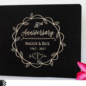 Puede incluir: Un &aacute;lbum de fotos de madera negra con encuadernaci&oacute;n en espiral. La portada presenta un dise&ntilde;o de corona dorada con las palabras "50th Anniversary", los nombres "Maggie & Rick" y los a&ntilde;os "1967 - 2017". Una flor rosa est&aacute; en la esquina inferior derecha.