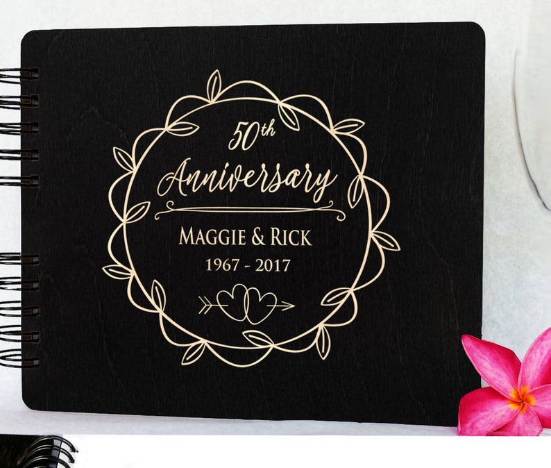 Puede incluir: Un &aacute;lbum de fotos de madera negra con encuadernaci&oacute;n en espiral. La portada presenta un dise&ntilde;o de corona dorada con las palabras "50th Anniversary", los nombres "Maggie & Rick" y los a&ntilde;os "1967 - 2017". Una flor rosa est&aacute; en la esquina inferior derecha.