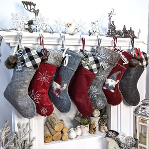 Puede incluir: Una colecci&oacute;n de calcetines navide&ntilde;os en varios colores y dise&ntilde;os, colgados de una repisa blanca. Los calcetines presentan patrones como cuadros de b&uacute;falo y bordados de copos de nieve, con etiquetas de nombre personalizadas. Algunos calcetines tienen dise&ntilde;os de gnomos y huellas de patas.