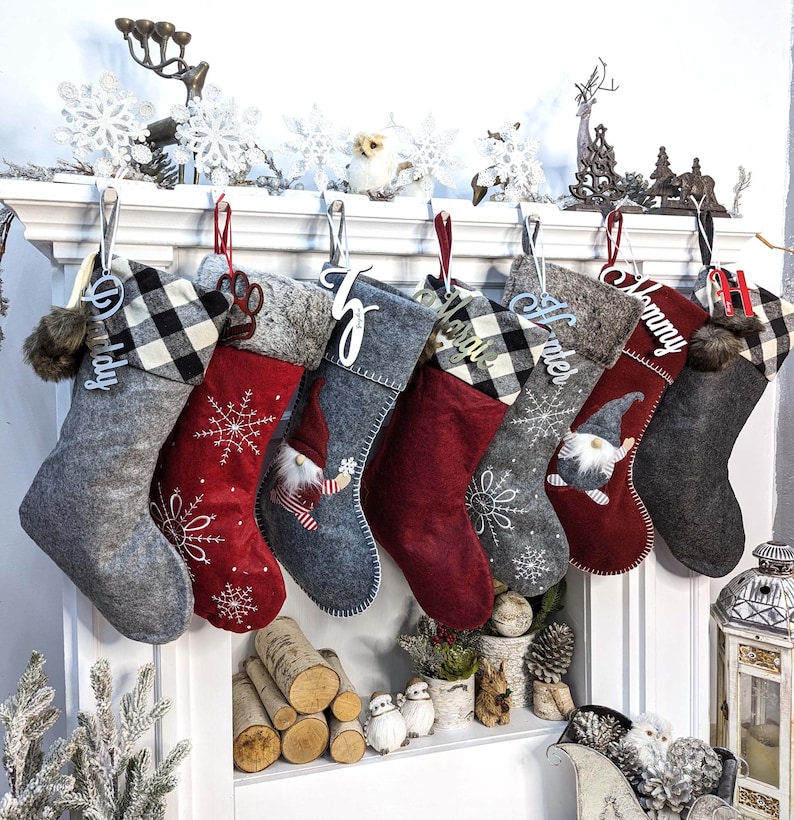 Puede incluir: Una colecci&oacute;n de calcetines navide&ntilde;os en varios colores y dise&ntilde;os, colgados de una repisa blanca. Los calcetines presentan patrones como cuadros de b&uacute;falo y bordados de copos de nieve, con etiquetas de nombre personalizadas. Algunos calcetines tienen dise&ntilde;os de gnomos y huellas de patas.
