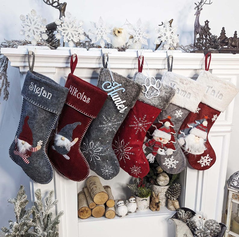 Puede incluir: Seis calcetines navide&ntilde;os decorativos, en tonos grises y rojos, personalizados con nombres como "Stefan" y "Nikolas". Presentan motivos festivos como copos de nieve y gnomos, colgados de una repisa blanca.