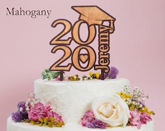 Pode incluir: Um topo de bolo de madeira para formatura com o ano "2020" e o nome "Jeremy". O topo apresenta um design de chapéu de formatura e um coração. O bolo é decorado com glacê branco e flores coloridas.