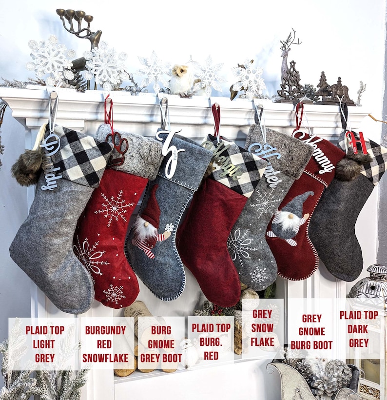 Puede incluir: Una colecci&oacute;n de calcetines navide&ntilde;os en varios colores y dise&ntilde;os, colgados de una repisa blanca. Los calcetines incluyen patrones de cuadros, copos de nieve y gnomos, con etiquetas de nombre personalizadas. Los colores incluyen gris, burdeos y rojo.
