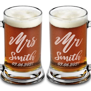 Pode incluir: Duas canecas de cerveja de vidro transparente cheias de cerveja e cobertas com espuma branca. Cada caneca tem uma al&ccedil;a e &eacute; gravada com "Mrs Smith" e "Mr Smith" em it&aacute;lico, juntamente com a data "03.06.2021".