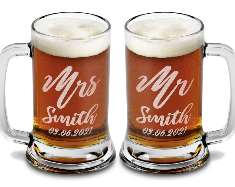 Pode incluir: Duas canecas de cerveja de vidro transparente cheias de cerveja e cobertas com espuma branca. Cada caneca tem uma al&ccedil;a e &eacute; gravada com "Mrs Smith" e "Mr Smith" em it&aacute;lico, juntamente com a data "03.06.2021".