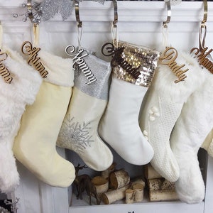 May include: Five white Christmas stockings with personalized wooden name tags. The stockings are decorated with fur, sequins, and snowflakes. The names on the tags are "Grandma", "Daddy", "Mommy", "Ariana", and "Giomo".