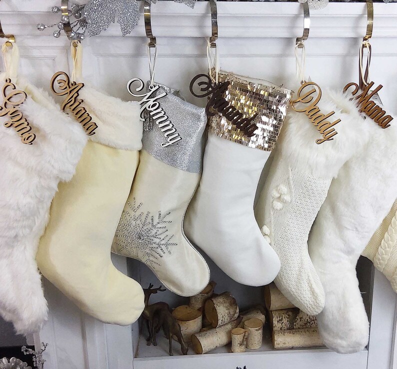 May include: Five white Christmas stockings with personalized wooden name tags. The stockings are decorated with fur, sequins, and snowflakes. The names on the tags are "Grandma", "Daddy", "Mommy", "Ariana", and "Giomo".