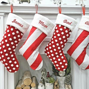 May include: Four Christmas stockings hang above a fireplace. Two are red with white polka dots, and two are red and white striped. Each stocking has a white cuff with a name embroidered in red: James, Mom, Dad, and Amber.