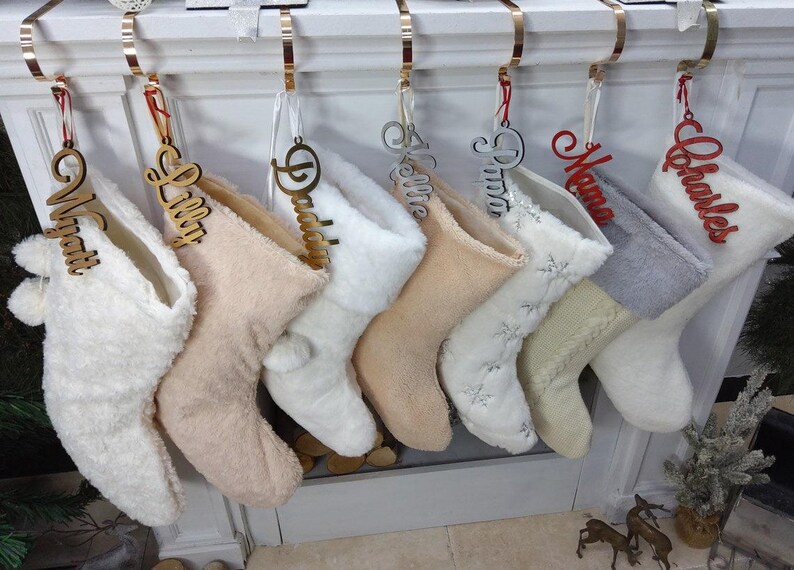 May include: Seven white and cream-colored stockings with gold and red personalized name tags. The names on the tags are "Wyatt", "Lilly", "Daddy", "Kellie", "Pizza", "Mama", and "Charles".