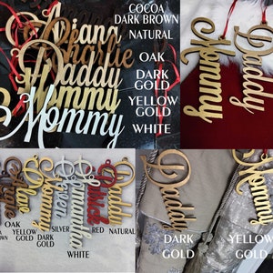 May include: A collection of personalized wooden ornaments in various colors, including natural, oak, dark gold, yellow gold, and white. The ornaments feature the words "Mommy", "Daddy", "Charlie", "Vivian", "Maggie", "Aubrey", "Patrick", and "Mommy".