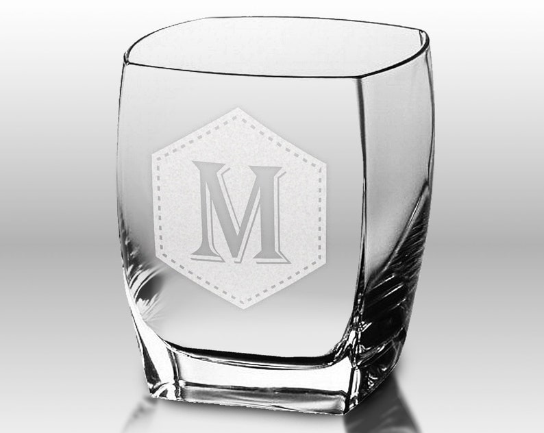Puede incluir: Un vaso transparente de forma cuadrada con un dise&ntilde;o curvo. El vaso presenta un dise&ntilde;o grabado de la letra "M" dentro de un hex&aacute;gono punteado. El vaso es adecuado para bebidas.