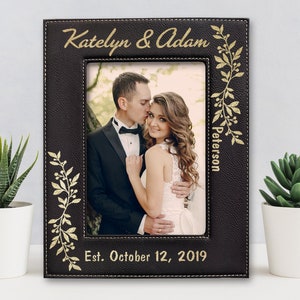 May include: Black leather photo frame with gold stitching and a gold floral design. The frame has a photo of a couple and the text "Katelyn & Adam Peterson Est. October 12, 2019".