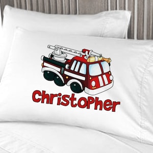 May include: White pillowcase featuring a cartoon fire truck in red, black, and yellow. The name "Christopher" is printed in red below the truck. The pillowcase is on a white bed.