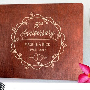 Puede incluir: Un &aacute;lbum de fotos de madera con encuadernaci&oacute;n en espiral. La portada est&aacute; grabada con las palabras "50th Anniversary", los nombres "Maggie & Rick" y los a&ntilde;os "1967-2017" dentro de una corona decorativa. Una flor rosa est&aacute; en la esquina.