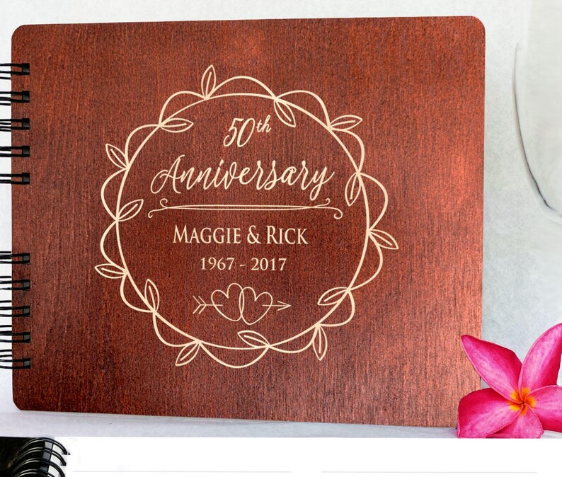 Puede incluir: Un &aacute;lbum de fotos de madera con encuadernaci&oacute;n en espiral. La portada est&aacute; grabada con las palabras "50th Anniversary", los nombres "Maggie & Rick" y los a&ntilde;os "1967-2017" dentro de una corona decorativa. Una flor rosa est&aacute; en la esquina.