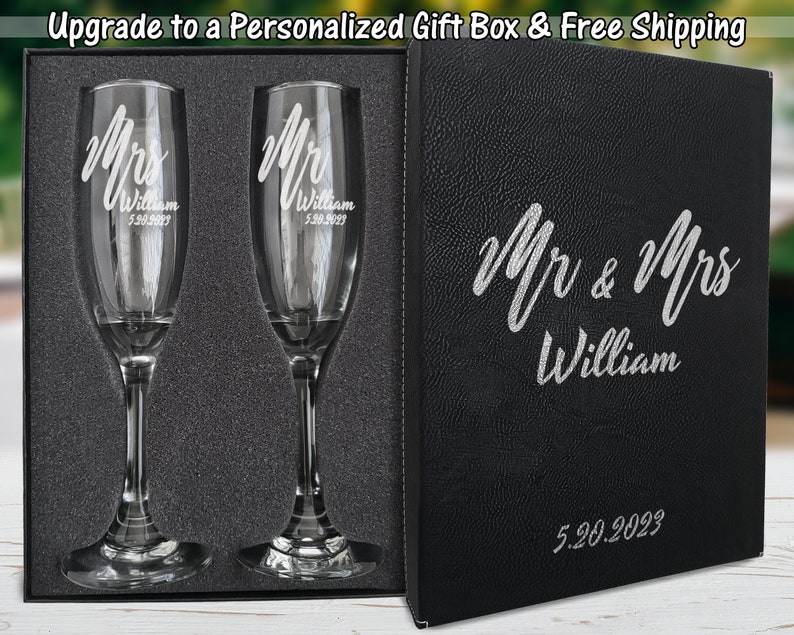 Pode incluir: Um conjunto de duas ta&ccedil;as de champanhe gravadas e uma caixa de presente preta. As ta&ccedil;as s&atilde;o gravadas com "Mr & Mrs William" e a data "5.20.2023". A caixa de presente tamb&eacute;m &eacute; personalizada com o mesmo texto e data.