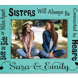 May include: A turquoise picture frame with a white stitched border and black text that reads "Side by Side or Miles Apart, Sisters Will Always Be Connected by Heart, Sara & Emily".
