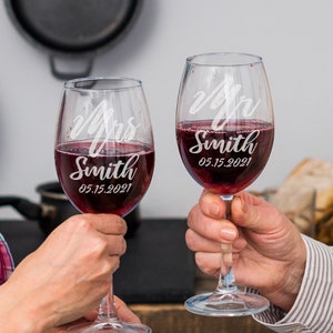 Pode incluir: Duas ta&ccedil;as de vinho transparentes cheias de vinho tinto. Cada ta&ccedil;a &eacute; gravada com as palavras "Mrs Smith" e "Mr Smith" juntamente com a data "05.15.2021". As ta&ccedil;as s&atilde;o levantadas, sugerindo um brinde.