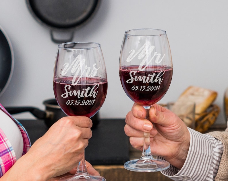 Pode incluir: Duas ta&ccedil;as de vinho transparentes cheias de vinho tinto. Cada ta&ccedil;a &eacute; gravada com as palavras "Mrs Smith" e "Mr Smith" juntamente com a data "05.15.2021". As ta&ccedil;as s&atilde;o levantadas, sugerindo um brinde.