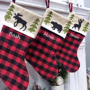 May include: Three red and black buffalo check Christmas stockings with personalised names "Bash", "Mom", and "Jensen". Each stocking features a burlap cuff with an embroidered design of a woodland scene with a moose, bear, and deer.
