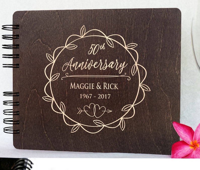 Puede incluir: Un &aacute;lbum de aniversario de madera marr&oacute;n oscuro con encuadernaci&oacute;n en espiral. La portada presenta el texto "50th Anniversary" dentro de una corona decorativa, junto con los nombres "Maggie & Rick" y los a&ntilde;os "1967-2017".