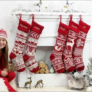 May include: Red and white Christmas stockings with personalized names hang above a fireplace. The stockings feature knitted patterns and are decorated with snowflakes, reindeer, and snowmen. A young girl in a matching red outfit and hat smiles in front of the fireplace.