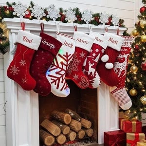 Könnte beinhalten: Ein dekorierter Kaminsims mit sechs roten Weihnachtsstrümpfen, jeweils mit weißem Bündchen und einem eingestickten Namen. Die Strümpfe sind mit Schneeflocken und anderen festlichen Designs verziert. Ein Weihnachtsbaum und verpackte Geschenke sind im Hintergrund.