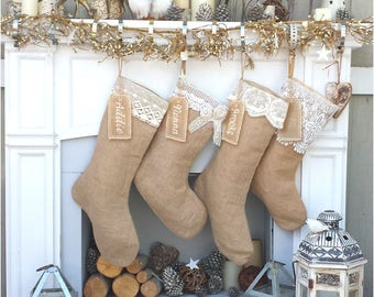 May include: Four burlap Christmas stockings with lace trim and personalized tags hanging on a white fireplace mantel. The tags read "Addie", "Hanna", "Brooke", and "_" . The stockings are decorated with a white bow and a white flower.
