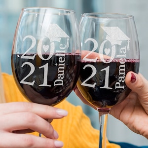 May include: Two clear wine glasses with red wine. The glasses are etched with the text "2021" and a graduation cap. One glass says "Danielle" and the other says "Pamela".
