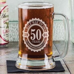 May include: A clear glass beer mug filled with amber-colored beer. The mug has a handle and a decorative emblem that reads "HALF WAY TO 100" with the number "50" in the center, and the years "1966" and "19" on either side. The mug sits on a black coaster.