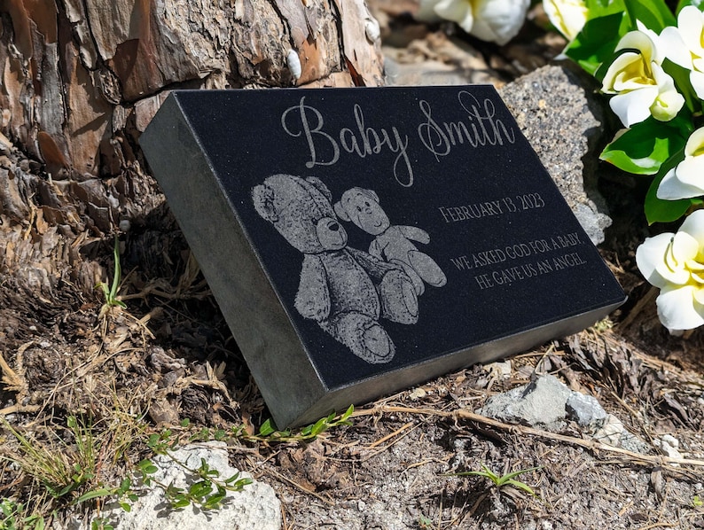 May include: Black granite memorial stone with the inscription "Baby Smith" and the date "February 13, 2023". The inscription continues with "We asked God for a baby, He gave us an angel". The stone is decorated with two teddy bears.