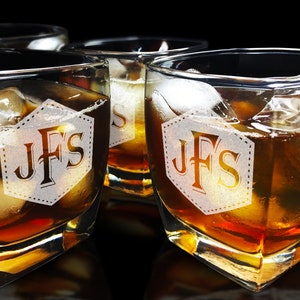 Puede incluir: Cuatro vasos cuadrados de whisky llenos de l&iacute;quido &aacute;mbar y cubitos de hielo. Cada vaso tiene las iniciales "JFS" grabadas dentro de un hex&aacute;gono blanco punteado. Los vasos est&aacute;n dispuestos sobre una superficie oscura.