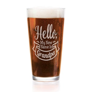 May include: A clear glass pint beer glass filled with amber liquid and white foam, featuring an etched white design that reads Hello, My New Name Is Grandpa, with the word Grandpa centered inside a decorative banner graphic element.