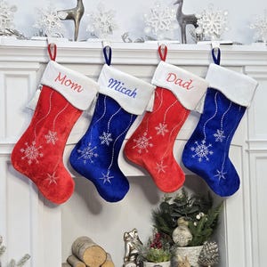 Könnte beinhalten: Vier personalisierte Weihnachtsstrümpfe mit Schneeflocken-Designs. Zwei Strümpfe sind aus rotem Samt mit weißen Schneeflocken und weißem Besatz. Zwei Strümpfe sind aus blauem Samt mit weißen Schneeflocken und weißem Besatz. Die Strümpfe sind mit "Mutter", "Micah", "Vater" und einem leeren Feld beschriftet.