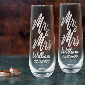 Pode incluir: Duas ta&ccedil;as de champanhe transparentes com as palavras "Mr & Mrs William 02.13.2020" gravadas. Duas alian&ccedil;as est&atilde;o em frente &agrave;s ta&ccedil;as. Um anel &eacute; dourado com uma banda prateada, o outro &eacute; prateado com uma banda de diamantes.
