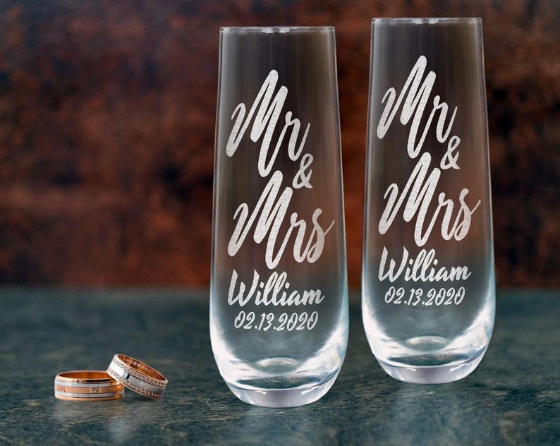 Pode incluir: Duas ta&ccedil;as de champanhe transparentes com as palavras "Mr & Mrs William 02.13.2020" gravadas. Duas alian&ccedil;as est&atilde;o em frente &agrave;s ta&ccedil;as. Um anel &eacute; dourado com uma banda prateada, o outro &eacute; prateado com uma banda de diamantes.