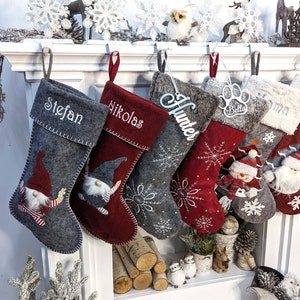 Puede incluir: Una colecci&oacute;n de calcetines navide&ntilde;os en varios tonos de gris y rojo, personalizados con nombres como "Stefan" y "Nikolas". Algunos calcetines presentan dise&ntilde;os de gnomos y patrones de copos de nieve, con acentos decorativos.