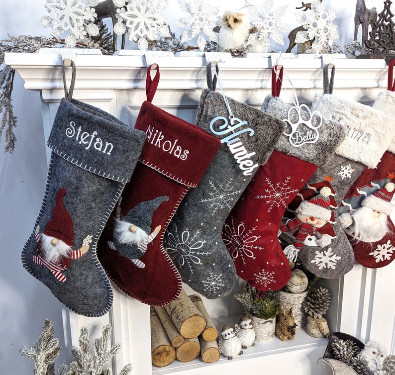 Puede incluir: Una colecci&oacute;n de calcetines navide&ntilde;os en varios tonos de gris y rojo, personalizados con nombres como "Stefan" y "Nikolas". Algunos calcetines presentan dise&ntilde;os de gnomos y patrones de copos de nieve, con acentos decorativos.