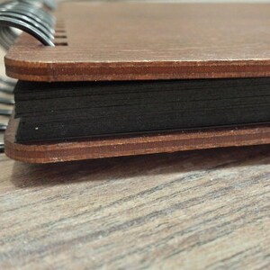 Puede incluir: Primer plano de un cuaderno con tapa de madera y p&aacute;ginas negras. La cubierta es de color marr&oacute;n c&aacute;lido, y las p&aacute;ginas est&aacute;n unidas por una espiral met&aacute;lica negra. El cuaderno descansa sobre una superficie de madera marr&oacute;n claro.