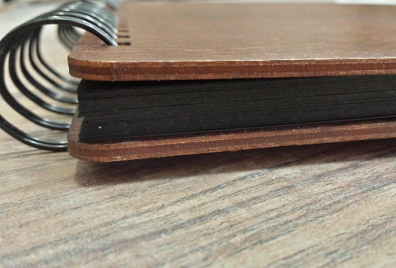 Puede incluir: Primer plano de un cuaderno con tapa de madera y p&aacute;ginas negras. La cubierta es de color marr&oacute;n c&aacute;lido, y las p&aacute;ginas est&aacute;n unidas por una espiral met&aacute;lica negra. El cuaderno descansa sobre una superficie de madera marr&oacute;n claro.