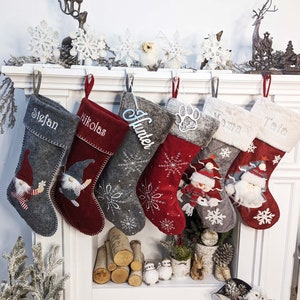 May include: A collection of Christmas stockings in various shades of gray and red, each personalized with names like "Stefan" and "Nikolas." The stockings feature festive designs including snowflakes, gnomes, and a snowman, with fluffy white trim.