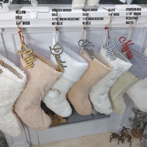 May include: Six personalized Christmas stockings with names "Man", "Lilly", "Daddy", "Kelle", "Papa", and "Charles" hanging on a white fireplace mantel. The stockings are made of white and beige faux fur and have pom-poms at the top.