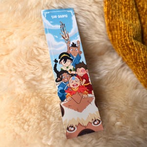 Peut inclure: Un marque-page rectangulaire en papier présentant une illustration colorée de personnages de dessin animé inspirés d'Avatar : Le Dernier Maître de l'Air. Le texte "the gaang" est imprimé en haut sur un fond de ciel bleu avec des nuages.